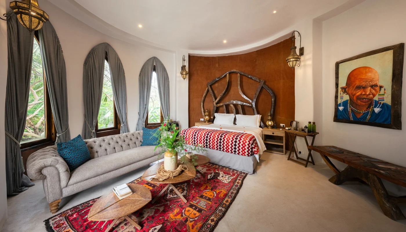 Luxurious upper floor bedroom at Mlima Villa in Xanadu Luxury Villas & Retreat, Zanzibar, featuring an artistic wooden headboard, a comfortable sofa, African artwork and tall arched windows.