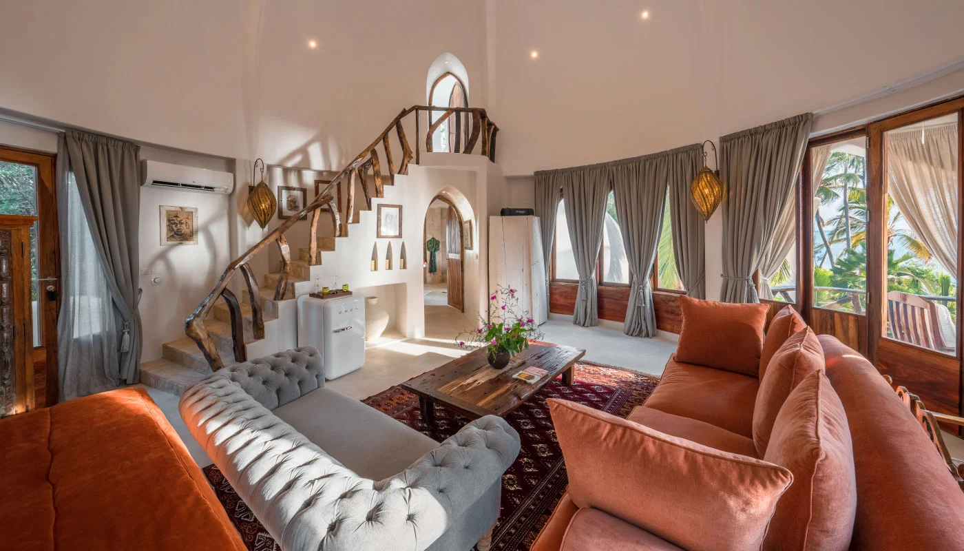Light-filled bedroom and living area on the second floor of Nyota Villa at Xanadu Luxury Villas & Retreat, Zanzibar, featuring comfortable sofas, a rustic wooden staircase and a balcony overlooking tropical palm trees.