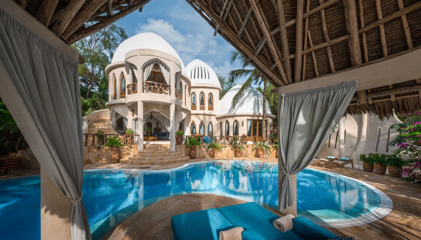 View from the covered lounge pavilion across the curved private pool towards the luxurious Nyota Villa at Xanadu Luxury Villas & Retreat, Zanzibar, featuring distinctive white domed architecture and lush tropical gardens.