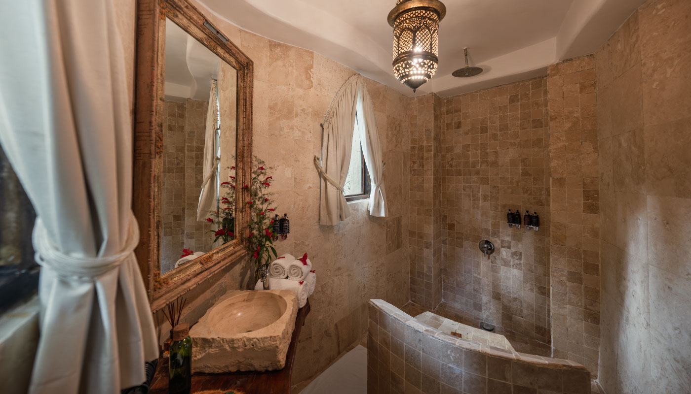Elegant stone bathroom in a luxury Zanzibar villa featuring a walk-in shower with love seat, handcrafted natural stone sink, Moroccan-style lantern lighting, and spa-inspired boutique resort design.