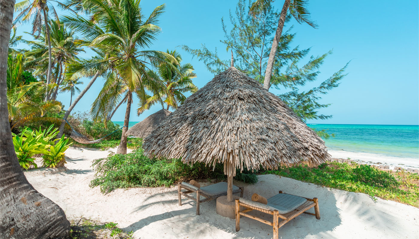 Tropical beachfront retreat in Zanzibar featuring traditional thatched sun loungers, white sand beach, turquoise Indian Ocean views, and palm trees surrounding a luxury boutique resort.