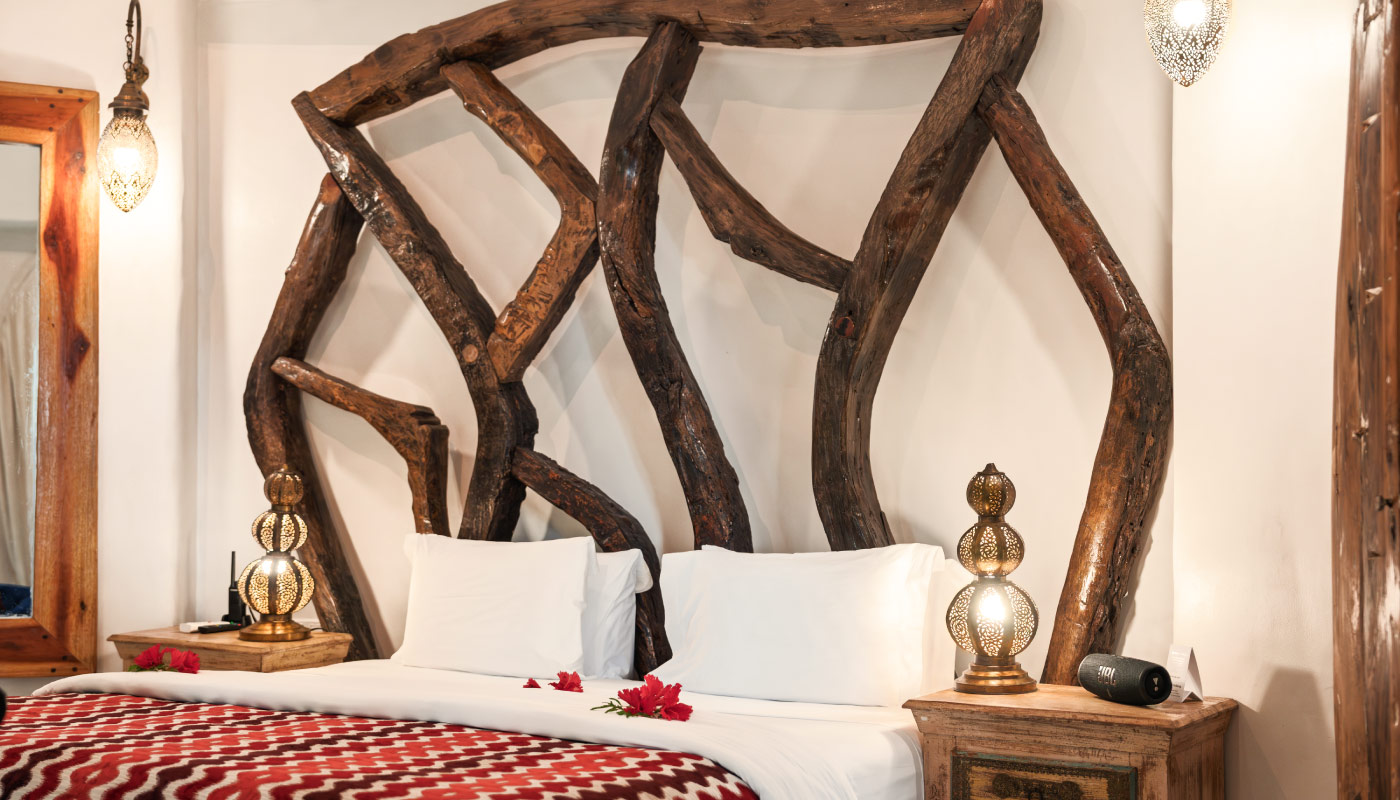 Luxury king-size bed with a handcrafted driftwood headboard, warm Moroccan lantern lighting, and elegant wooden décor in a boutique Zanzibar villa suite.