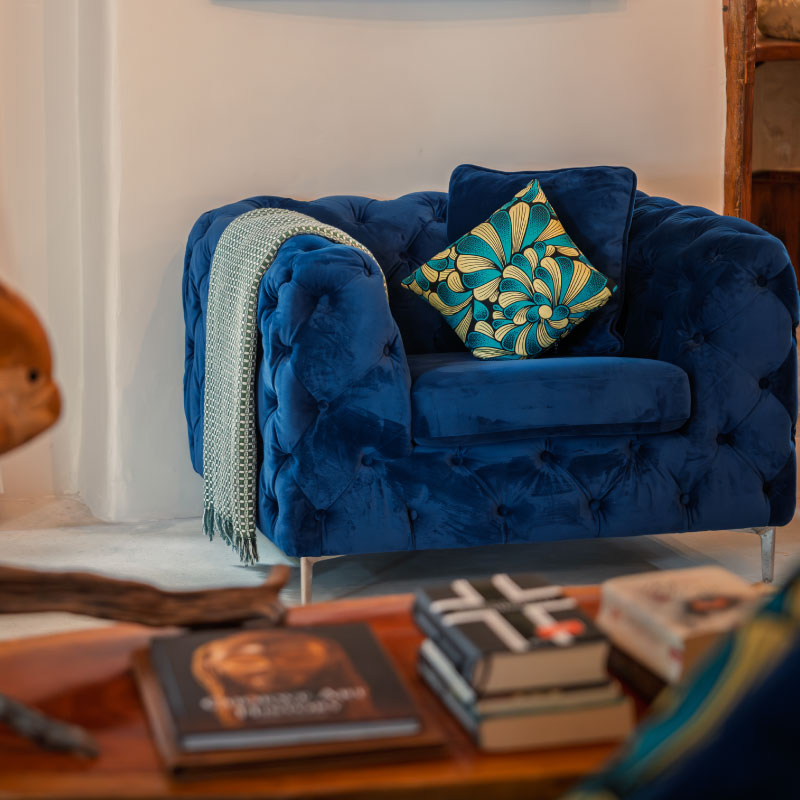 Blue velvet armchair with decorative kitenge cushions in a luxury Zanzibar villa suite, featuring books and handcrafted wooden furnishings.